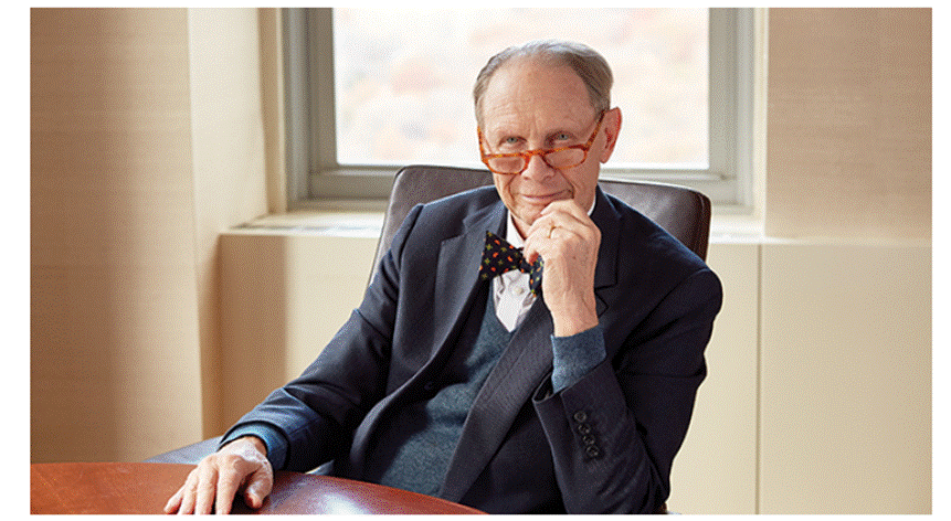 A person in a suit and bow tie sitting at a table
Description automatically generated